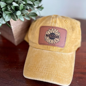 May include: A yellow canvas baseball cap with a brown leather patch featuring a sunflower and the word "mama" in black lettering.