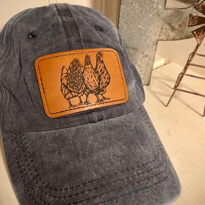 May include: A dark blue denim baseball cap with a brown leather patch featuring a design of three chickens.