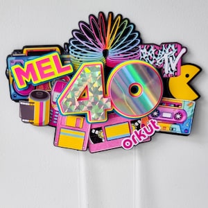 May include: A vibrant 80s-themed cake topper featuring the name "MEL" and the number "40". The design incorporates a rainbow slinky, a CD, a Pac-Man, a cassette tape, and the MTV logo. The topper is composed of various shapes and colours.