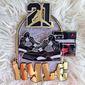 May include: A gold and black cake topper featuring the number 21, a basketball player silhouette, a pair of black and white trainers, and the name "KYLE" in gold lettering.