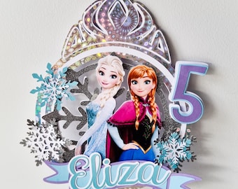 May include: A purple and silver cake topper featuring the characters Elsa and Anna from the movie Frozen. The topper includes a crown, snowflakes, and the number 5. The name "Eliza" is written in a blue banner.