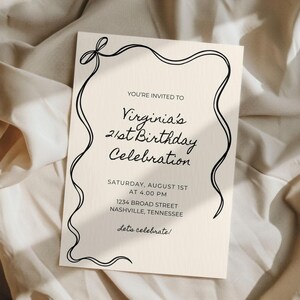 Puede incluir: Una tarjeta de invitaci&oacute;n crema con texto negro. El texto dice "Virginia's 21st Birthday Celebration" con la fecha, hora y direcci&oacute;n. Un dise&ntilde;o de cinta negra enmarca el texto, agregando un toque decorativo.