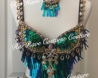 May include: A green and gold sequined bra top with fringe and a matching skirt. The bra top has a large, ornate pendant with rhinestones and gold accents. The skirt is also adorned with fringe and rhinestones. The text "Rio Rave Costume Couture" is visible on the image.