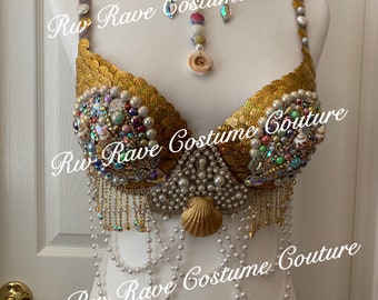May include: Gold and pearl embellished bra top with a seashell design. The bra top has a gold necklace with pearls and iridescent stones. The text "Ru Rave Costume Couture" is visible on the image.