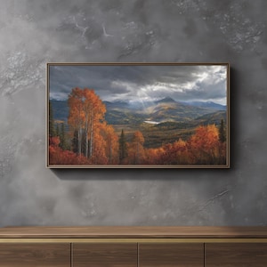 May include: A rectangular framed landscape wall art print featuring a mountain range under a cloudy sky with sunbeams. The foreground displays vibrant orange and red autumn trees, while the background shows a valley with a small lake.