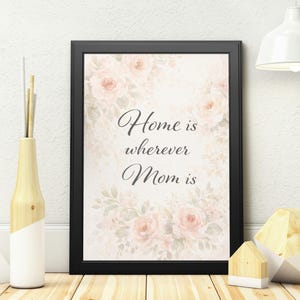 Könnte beinhalten: Gerahmter Druck mit dem Text "Home is wherever Mom is" in eleganter Schrift. Das Kunstwerk zeigt einen weichen floralen Hintergrund mit rosa Rosen und grünen Blättern. Der Rahmen ist schwarz, und der Druck befindet sich auf einer Holzoberfläche.