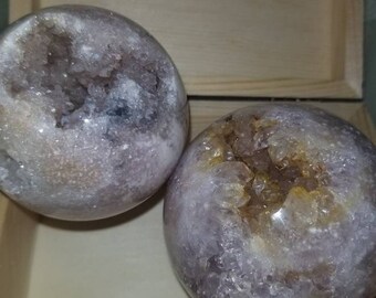 May include: Two amethyst crystal spheres with a purple and white color scheme. The spheres are in a wooden box and have a natural, rough texture.