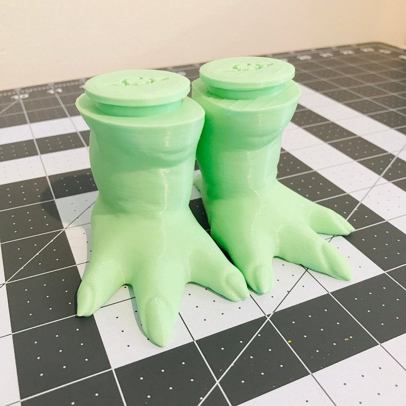 May include: A pair of green 3D printed Yoda feet with a textured surface. The feet are designed to be used as bottle stoppers.