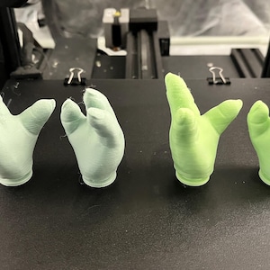 May include: Four small, 3D-printed hand sculptures in varying shades of green. Two are a light mint green, and two are a brighter, almost neon green. The hands are posed in different gestures, showcasing the detail of the 3D printing process.