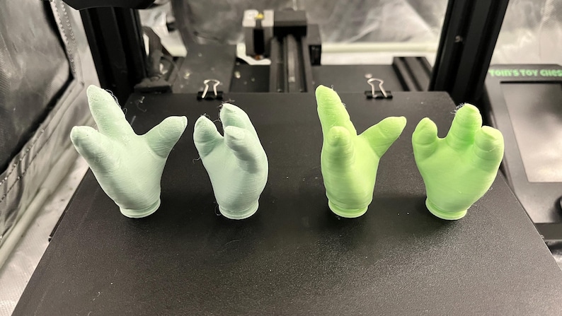 May include: Four small, 3D-printed hand sculptures in varying shades of green. Two are a light mint green, and two are a brighter, almost neon green. The hands are posed in different gestures, showcasing the detail of the 3D printing process.