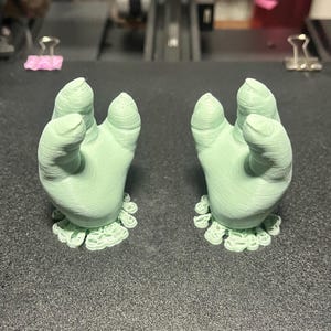 May include: Two light green, 3D-printed hand sculptures with open palms and detailed finger and wrist features. Each hand rests on a decorative, ruffled base. The hands are positioned on a dark gray surface.