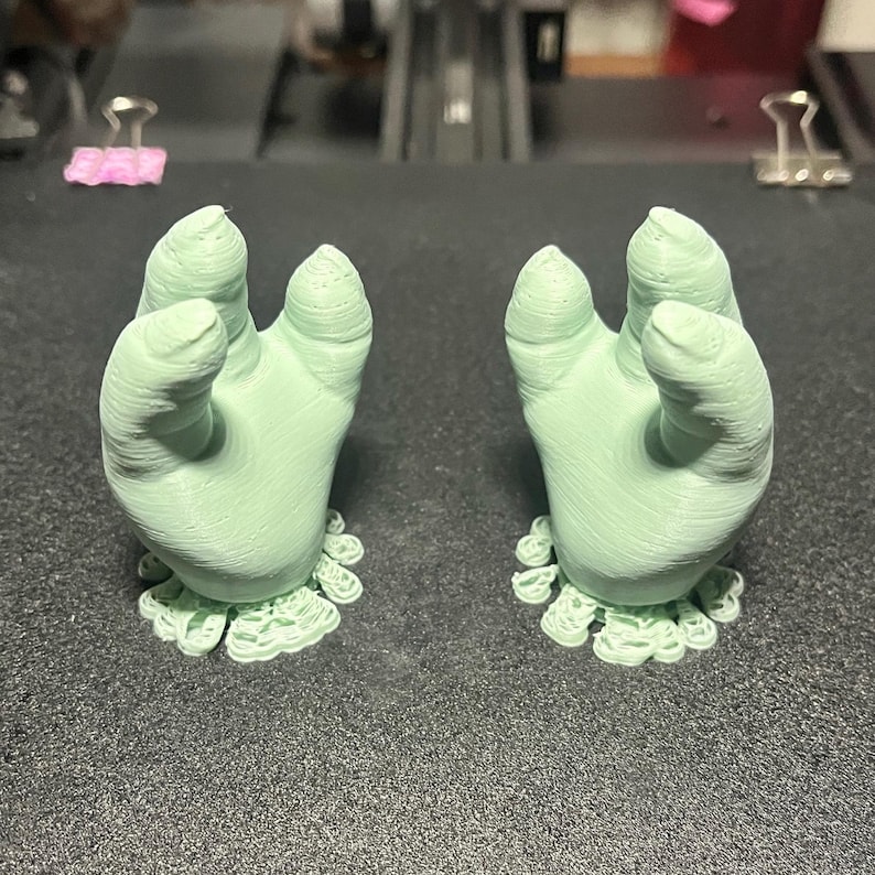 May include: Two light green, 3D-printed hand sculptures with open palms and detailed finger and wrist features. Each hand rests on a decorative, ruffled base. The hands are positioned on a dark gray surface.