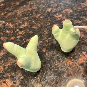 May include: Two light green, hand-shaped figurines with tan-colored fingertips. One hand is in a peace sign gesture, and the other is open. The figurines are on a dark, speckled surface.