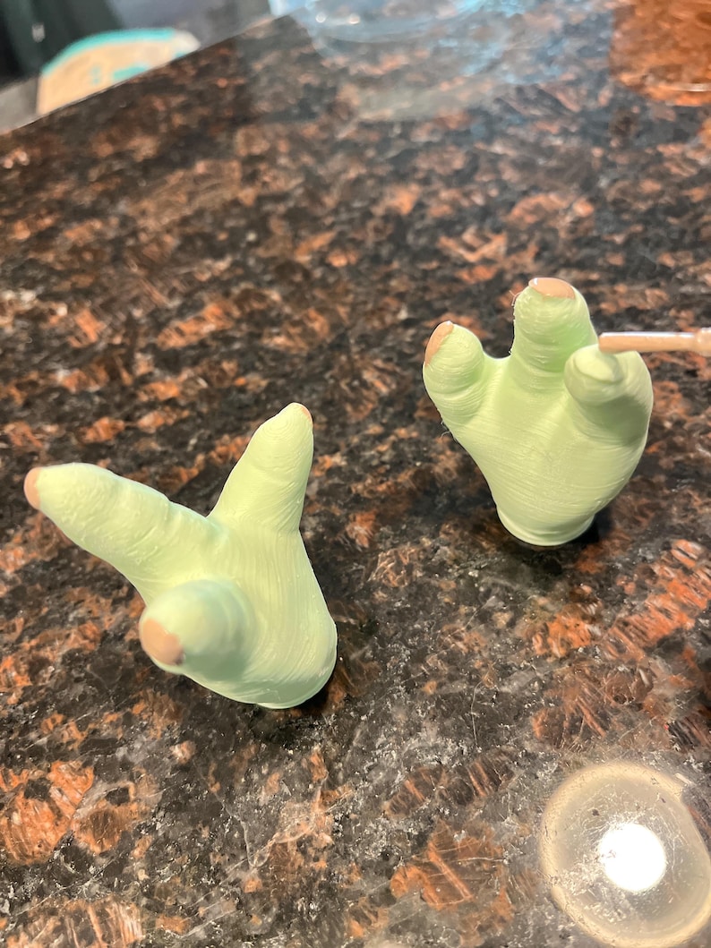 May include: Two light green, hand-shaped figurines with tan-colored fingertips. One hand is in a peace sign gesture, and the other is open. The figurines are on a dark, speckled surface.