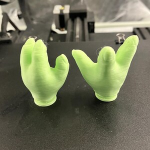 May include: Two light green, 3D-printed hand sculptures, each with a palm and four fingers. The hands are positioned on a black surface, likely a printer bed. The sculptures are detailed and appear to be freshly printed.