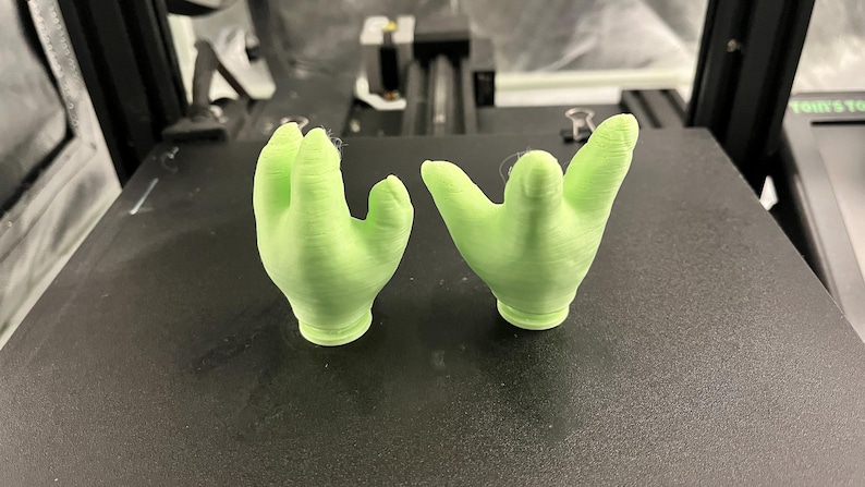 May include: Two light green, 3D-printed hand sculptures, each with a palm and four fingers. The hands are positioned on a black surface, likely a printer bed. The sculptures are detailed and appear to be freshly printed.