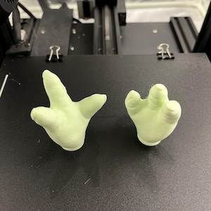 May include: Two light green, 3D-printed hand models. One hand is open, and the other is closed. The hands are resting on a black surface, likely a 3D printer bed. The hands are a pale, almost pastel green color.