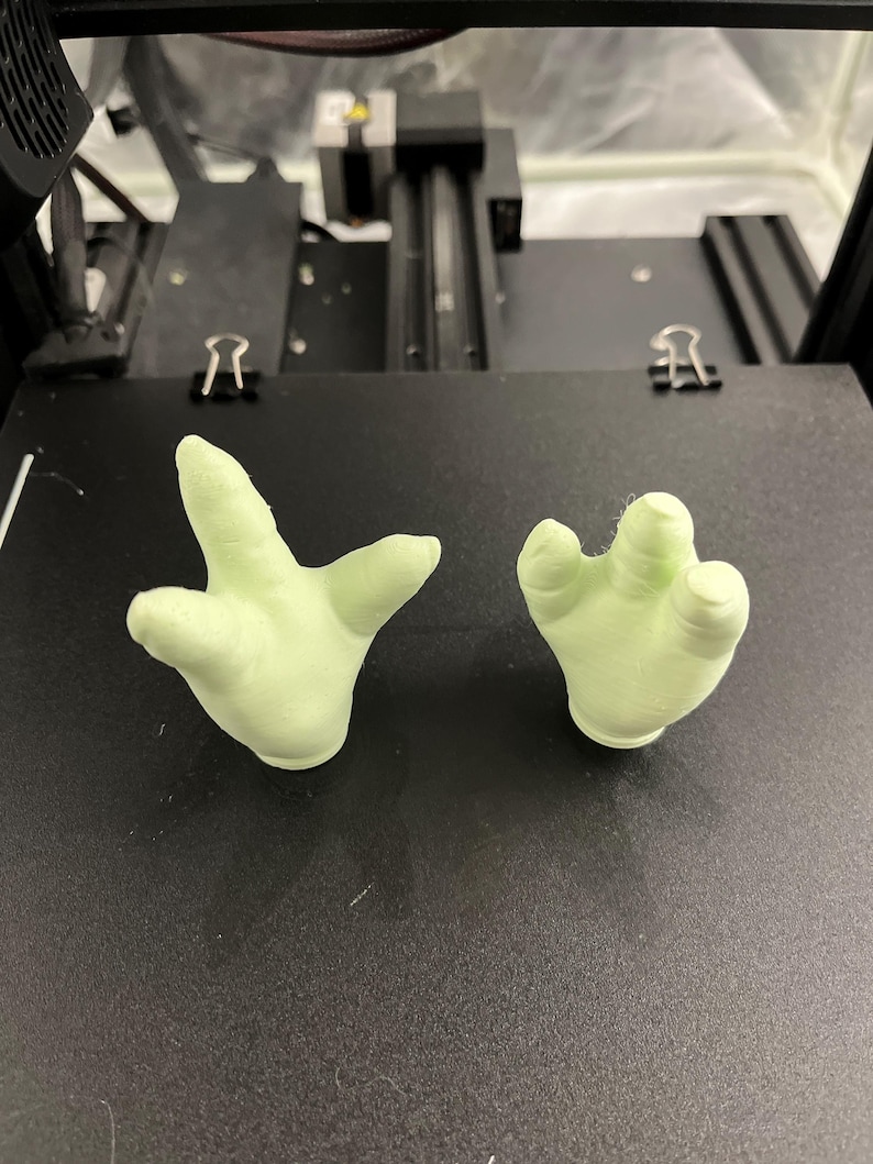 May include: Two light green, 3D-printed hand models. One hand is open, and the other is closed. The hands are resting on a black surface, likely a 3D printer bed. The hands are a pale, almost pastel green color.