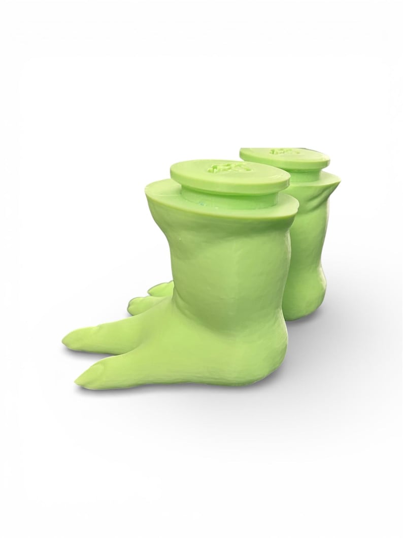 May include: Two light green, foot-shaped bottle stoppers. Each stopper has a flat, circular top and a curved, boot-like base with toes. The stoppers are made of a smooth material and are set against a white background.