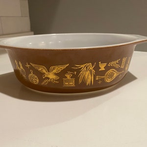 Puede incluir: Una fuente de horno marrón con interior blanco y borde dorado. La fuente tiene un patrón de imágenes doradas alrededor del borde, incluyendo un pájaro, una mazorca de maíz, un mortero y un pilón, y un abanico.