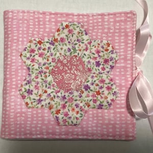 May include: A pink fabric book with a white polka dot pattern and a pink ribbon. The front cover features a large flower made of fabric with a floral pattern.
