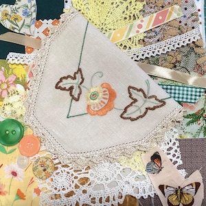 May include: A collection of vintage fabric scraps, featuring an embroidered cream handkerchief with orange and green floral motifs. Other elements include lace, buttons, ribbons, and butterfly appliqués. The colour scheme is soft, with greens, yellows, and oranges.