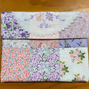 May include: A rectangular fabric pouch featuring a patchwork design of various floral patterns in shades of purple, pink, and green. The top flap is white with embroidered pink and purple flowers and a white lace trim along the curved edge.