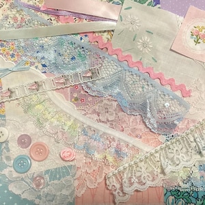 May include: A collection of pastel-coloured fabric scraps, ribbons, and lace. Features light blue and pink lace, floral patterned fabric, and various buttons. A small pink rose and a light blue bow are also visible.