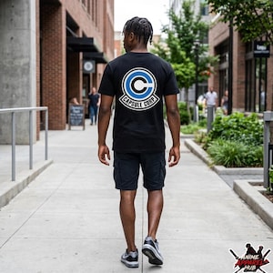 May include: Black t-shirt with a Capsule Corp logo on the back, featuring a blue and white design. The person is wearing black shorts and gray sneakers, walking on a sidewalk.