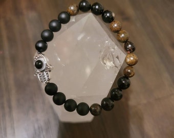 May include: A beaded bracelet featuring matte black and brown gemstone beads. The bracelet includes a silver-colored Hamsa hand charm with an eye design. The bracelet is displayed on a clear, faceted surface.
