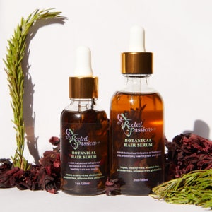 May include: Two glass bottles of hair serum with gold caps and labels that say "Rooted Passion+ Botanical Hair Serum". The smaller bottle is 1 oz/30 ml and the larger bottle is 2 oz/60 ml. Both bottles are sitting on a surface with dried red flowers and green rosemary sprigs.