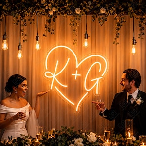 May include: A heart-shaped neon sign displaying the letters K and G with a plus sign in the center, emitting a warm orange glow. The sign is mounted against a draped fabric backdrop illuminated by hanging Edison bulbs and greenery.