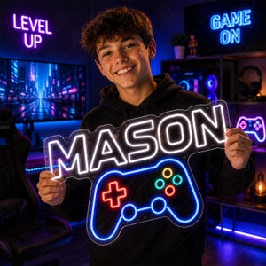 May include: A custom neon sign featuring the name MASON in bright white capital letters positioned above a blue neon outline of a video game controller with red and green button accents, held against a dark background with gaming decor.