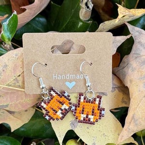 May include: A pair of handmade earrings featuring a pixelated fox design in orange, brown, and white. The earrings are on a card with the word "Handmade" and a heart.