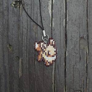 May include: A beaded pendant necklace featuring a pixelated design in shades of brown, cream, and red. The pendant is attached to a black cord with a silver-colored clasp. The necklace is displayed on a weathered wooden surface.