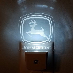 Peut inclure: Une veilleuse avec le logo John Deere blanc branchée sur une prise électrique. Le logo représente une silhouette de cerf blanche sur fond vert.