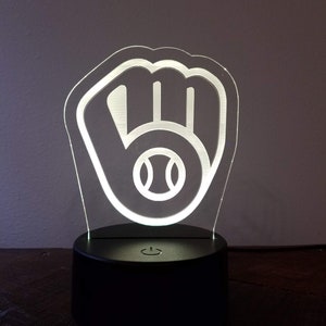 Pode incluir: Uma luz noturna de acrílico branco em forma de luva de beisebol com uma bola de beisebol dentro. A luz noturna está sobre uma base preta com um interruptor de ligar/desligar.