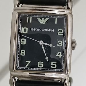 May include: A rectangular Emporio Armani wristwatch with a black face and silver case. The watch has white numerals, hands, and the brand's logo. The black leather strap is attached to the case.