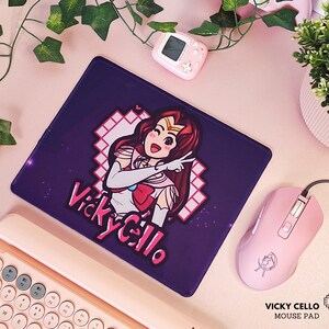 Może przedstawiać: Fioletowa podkładka pod mysz z różową i białą postacią anime w stroju marynarskim. Postać pokazuje znak pokoju, a tekst "Vicky Cello" jest napisany różowymi i białymi literami.
