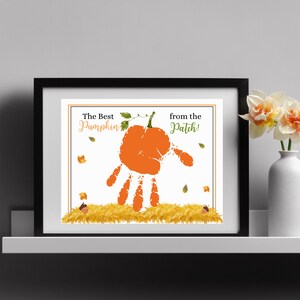Könnte beinhalten: Ein gerahmter Druck mit einem Handabdruck in Form eines Kürbisses mit einem grünen Stiel und Blättern. Der Text lautet "The Best Pumpkin from the Patch!" mit Herbstblättern und Eicheln, die um den Kürbis herum verstreut sind.