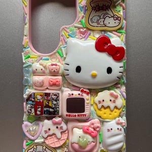 May include: A decorative phone case featuring a pink interior and a colorful exterior adorned with various Hello Kitty-themed embellishments. The case includes a large Hello Kitty face with a red bow, along with other charms and sprinkles.