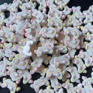 May include: A collection of white, decorative game controller-shaped embellishments. Each controller features pink, yellow, and blue floral and dot designs. The embellishments are scattered across a dark surface.