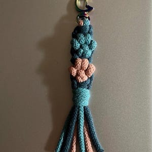 May include: A macrame keychain featuring a moon-shaped clasp. The keychain has a teal and tan color scheme, with a series of knotted flower-like shapes and a tassel at the bottom. The keychain is handmade and has a unique, artistic design.