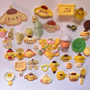 Könnte beinhalten: Eine Sammlung von Pompompurin-Harz-Cabochons und Anhängern in verschiedenen Formen, darunter Figuren, Lebensmittel und Zubehör. Die Teile sind in Gelb-, Braun- und Pastelltönen gehalten und zeigen teilweise kleine Aufdrucke und Symbole.