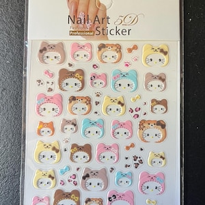 Könnte beinhalten: Ein Bogen mit 5D-Nagelaufklebern mit verschiedenen Katzenmotiven. Die Aufkleber zeigen zahlreiche kleine Katzengesichter mit Kapuzen in Farben wie Rosa, Blau, Braun und Gelb sowie winzige Schleifen und Pfotenabdrücke. Die Überschrift lautet Nail Art 5D Sticker.