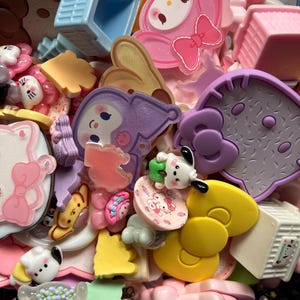 May include: A collection of colorful plastic charms featuring various cartoon characters, including pink and white cat-like figures, purple rabbit-like shapes, yellow bows, and small miniature furniture pieces in pastel shades of blue, pink, and white.