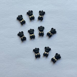 May include: A collection of nine small, matte black bear-shaped charms. Each bear features a small, shiny gold-colored crown on its head and a matching gold-colored bow tie detail at the neck, arranged in a scattered pattern on a plain surface.