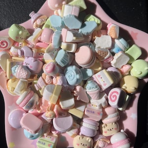 以下が含まれることがあります： パステルカラーのミニチュアフードチャームのコレクション。マカロン、アイスクリームコーン、ケーキ、クマの形をしたキャンディーなど、ピンクの星型の皿に並べられています。チャームはピンク、ブルー、イエロー、グリーンの色合いで、「milk」の文字が入っているものもあります。