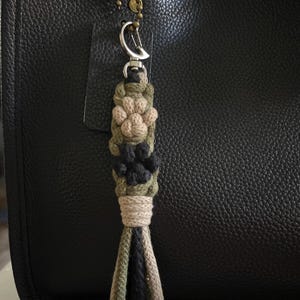 Op de afbeelding: Een handgemaakte macramé sleutelhanger met een hondenpootafdruk ontwerp. De sleutelhanger heeft een combinatie van groene, beige en zwarte kleuren. Het is bevestigd aan een zwarte leren tas met een goudkleurige ketting.