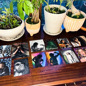 May include: A collection of 12 small square cards with various illustrations. The cards feature black and white drawings of people, a green monster, and a person in a white robe. The cards are arranged on a wooden table.